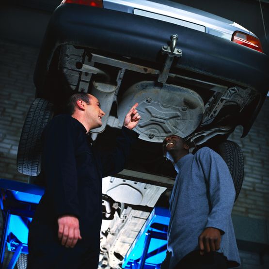 portrait of a young man standing below the undercarriage of his car with a mechanic